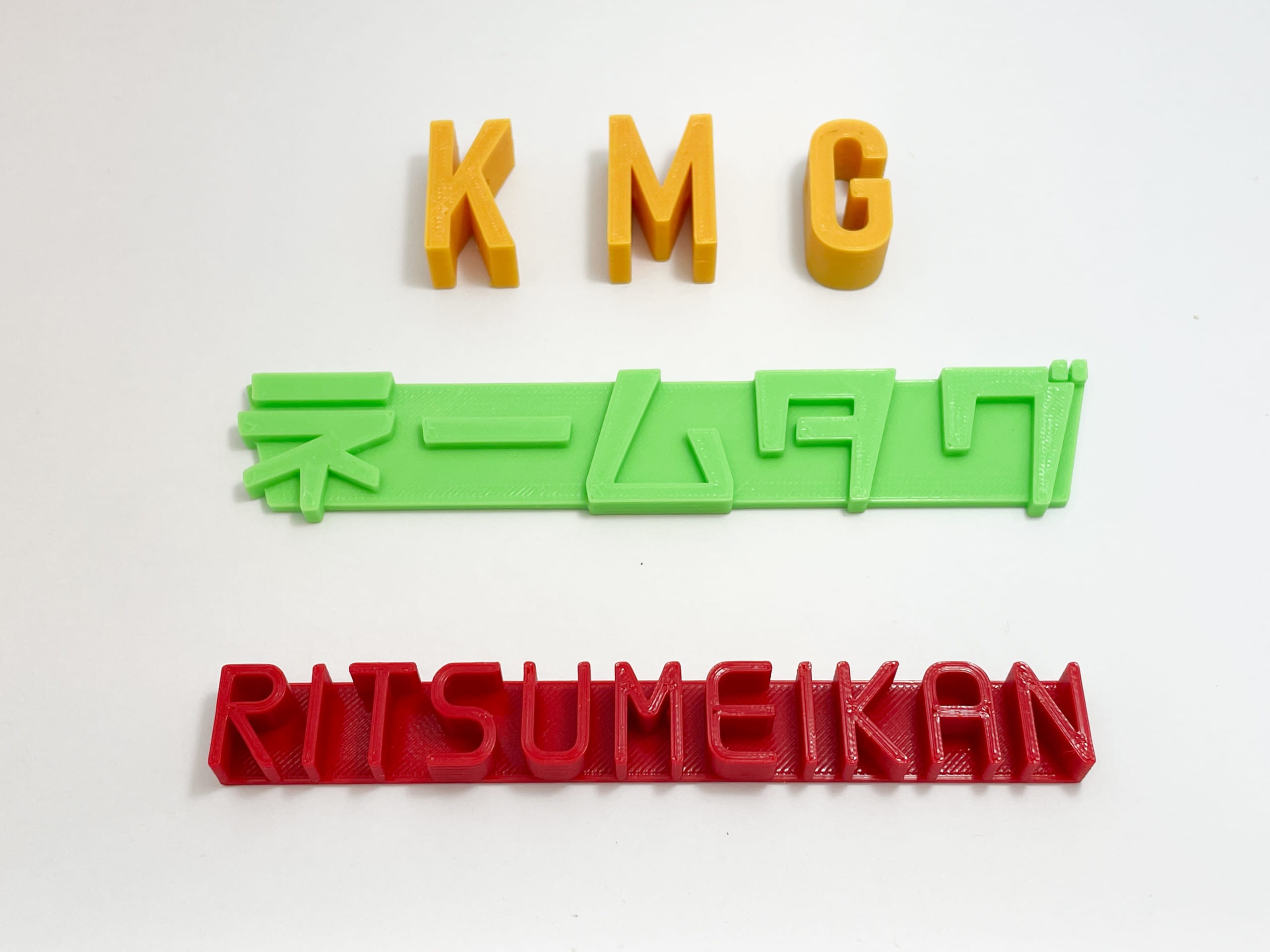 白い背景に3Dプリントされて文字が立体的になったネームタグがある。上は黄色い文字でKMG、真ん中は緑で「ネームタグ」、下は赤でRITSUMEIKAN