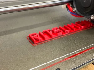 3Dプリント中のネームタグ。赤いフィラメントでRITSUMEIKANの文字が3Dプリンタの上でプリントされている